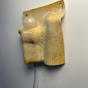 May include: A wall-mounted ceramic light fixture with a yellow glaze and dark brown speckles. The piece has an irregular, folded, and organic shape with a circular opening holding a round, frosted white glass light bulb and a thin power cord.