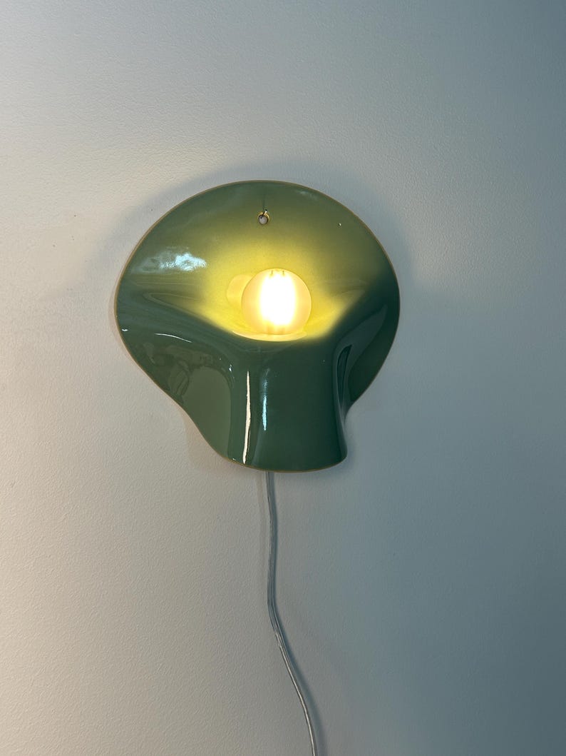 May include: A wall-mounted ceramic lamp with a glossy sage green finish and an organic, curved shape. The fixture features a central circular opening that houses a glowing, warm-toned light bulb, with a thin white power cord extending downward.
