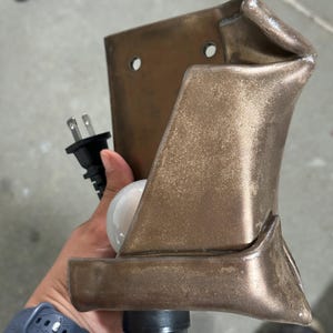 May include: A wall-mounted lamp fixture with a metallic bronze finish and a folded, sculptural design. The fixture features two circular mounting holes at the top, a black electrical cord with a standard plug, and a white light bulb socket.