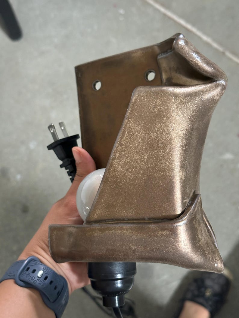 May include: A wall-mounted lamp fixture with a metallic bronze finish and a folded, sculptural design. The fixture features two circular mounting holes at the top, a black electrical cord with a standard plug, and a white light bulb socket.