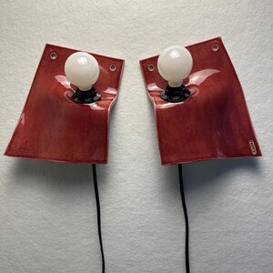 Puede incluir: Un par de l&aacute;mparas de pared con placas de cer&aacute;mica roja brillante y curvas suaves. Cada l&aacute;mpara cuenta con una bombilla esf&eacute;rica blanca centrada en la placa, cables el&eacute;ctricos negros que se extienden hacia abajo y orificios de montaje.