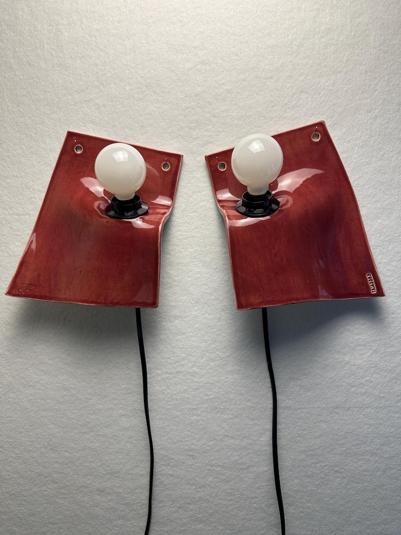 Puede incluir: Un par de l&aacute;mparas de pared con placas de cer&aacute;mica roja brillante y curvas suaves. Cada l&aacute;mpara cuenta con una bombilla esf&eacute;rica blanca centrada en la placa, cables el&eacute;ctricos negros que se extienden hacia abajo y orificios de montaje.
