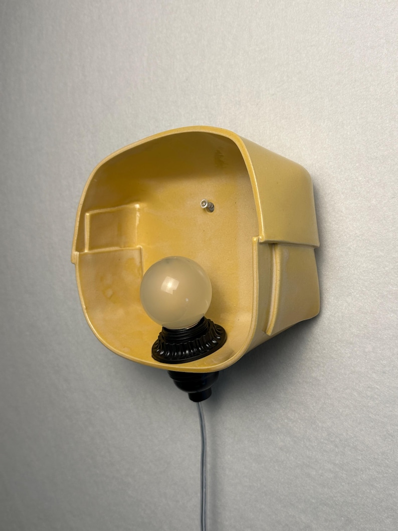 May include: A wall-mounted lamp featuring a smooth, yellow ceramic shade with an asymmetrical, rounded rectangular shape. A black socket holds a single, frosted white globe light bulb, and a thin gray power cord extends from the bottom.