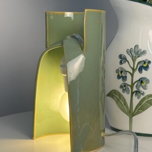 May include: A sage green ceramic table lamp with a curved, open-sided cylindrical structure. The lamp features a smooth, glossy finish and houses a single glowing light bulb inside the hollow center. A clear power cord extends from the base.