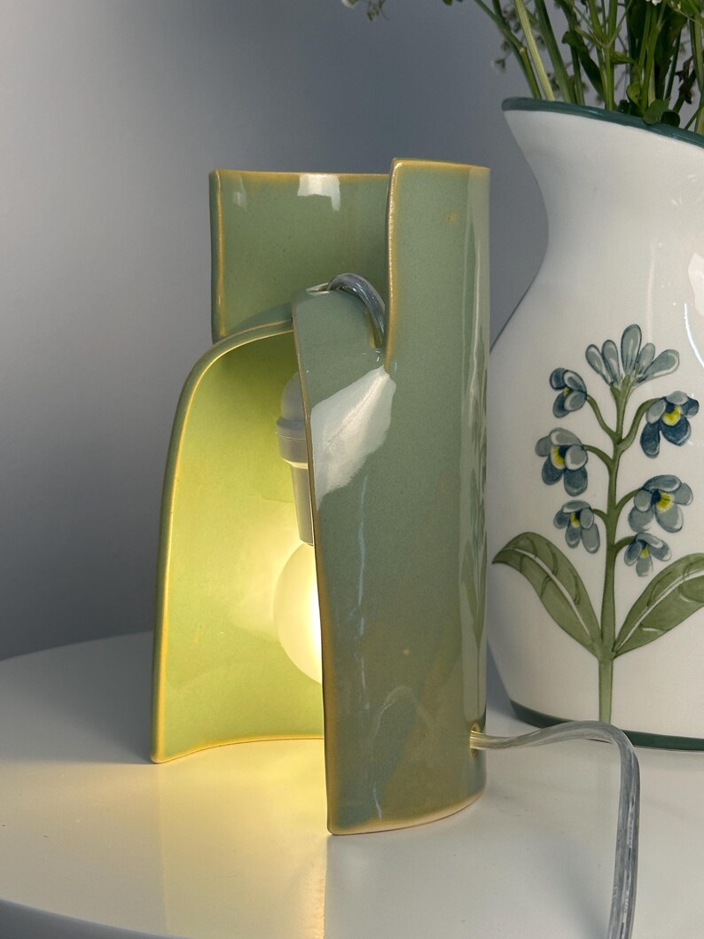May include: A sage green ceramic table lamp with a curved, open-sided cylindrical structure. The lamp features a smooth, glossy finish and houses a single glowing light bulb inside the hollow center. A clear power cord extends from the base.