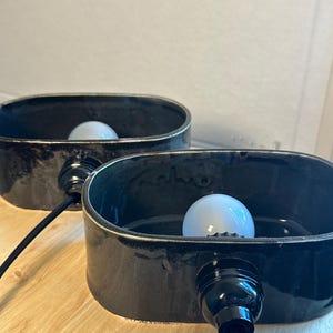 May include: Two black ceramic lamps with oval shades. Each lamp has a white light bulb and a black cord. The lamps are on a light-coloured wooden surface. The lamps are a modern home decor item.