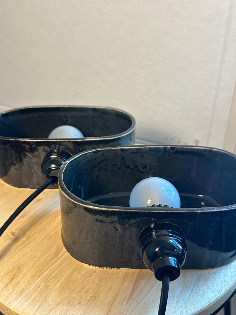 May include: Two black ceramic lamps with oval shades. Each lamp has a white light bulb and a black cord. The lamps are on a light-coloured wooden surface. The lamps are a modern home decor item.