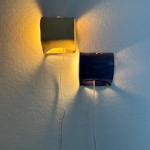 May include: Two wall-mounted sconces with curved, rectangular shades. One shade is a matte yellow emitting a warm glow, while the other is a dark navy blue. Each fixture features a thin, clear power cord hanging downward from the center base.