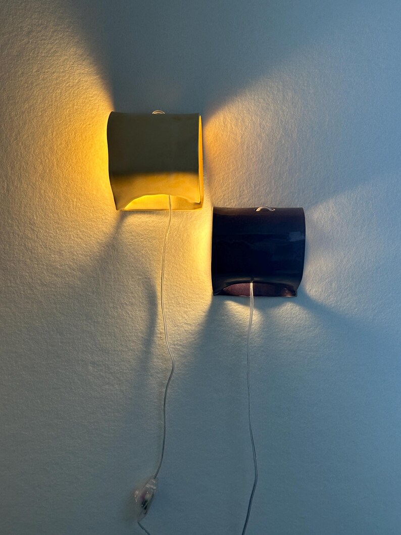 May include: Two wall-mounted sconces with curved, rectangular shades. One shade is a matte yellow emitting a warm glow, while the other is a dark navy blue. Each fixture features a thin, clear power cord hanging downward from the center base.