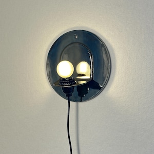 May include: A circular, dark-colored wall sconce with a glossy finish featuring a small, protruding shelf that holds a single, glowing spherical light bulb. A thin black power cord extends downward from the base of the light fixture.