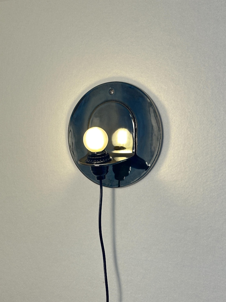 May include: A circular, dark-colored wall sconce with a glossy finish featuring a small, protruding shelf that holds a single, glowing spherical light bulb. A thin black power cord extends downward from the base of the light fixture.