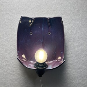 May include: A wall-mounted ceramic lamp featuring a glossy, deep purple and blue gradient finish. The curved, rectangular shade has two small circular holes near the top and a central socket holding a round, frosted white light bulb.