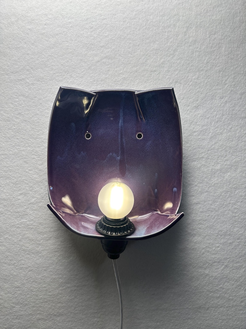 May include: A wall-mounted ceramic lamp featuring a glossy, deep purple and blue gradient finish. The curved, rectangular shade has two small circular holes near the top and a central socket holding a round, frosted white light bulb.