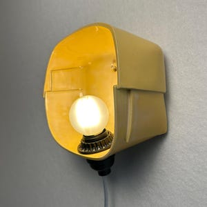 May include: A wall-mounted lamp featuring a pale yellow, curved, box-like shade with a smooth finish. A single, round, frosted white light bulb is centered within the shade, attached to a dark base with a thin, light-colored electrical cord.