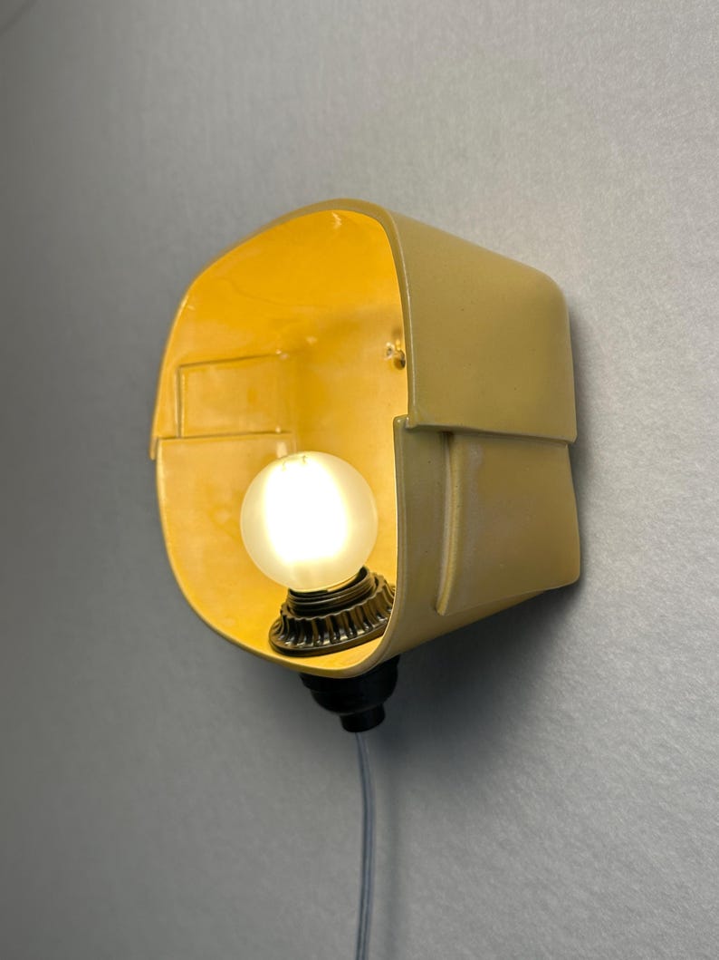 May include: A wall-mounted lamp featuring a pale yellow, curved, box-like shade with a smooth finish. A single, round, frosted white light bulb is centered within the shade, attached to a dark base with a thin, light-colored electrical cord.