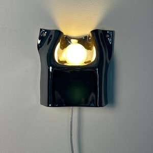May include: A glossy black wall-mounted lamp with a fluid, organic shape and a central cutout. A single round, warm-toned light bulb is positioned within the recessed center, casting a soft glow upward. A thin, clear power cord hangs from the bottom.
