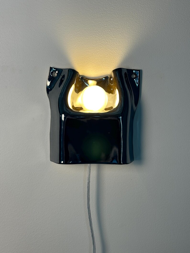 May include: A glossy black wall-mounted lamp with a fluid, organic shape and a central cutout. A single round, warm-toned light bulb is positioned within the recessed center, casting a soft glow upward. A thin, clear power cord hangs from the bottom.