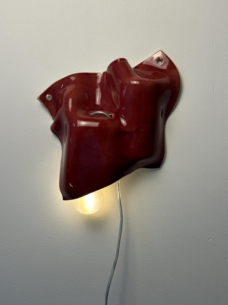 Puede incluir: Una l&aacute;mpara de pared con una forma cer&aacute;mica org&aacute;nica de color rojo intenso brillante y contornos irregulares. Una bombilla redonda y brillante sobresale del borde inferior, y un cable de alimentaci&oacute;n fino y transparente cuelga verticalmente.