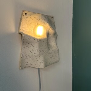 May include: A wall-mounted ceramic lamp with an off-white speckled finish and an irregular, wavy surface. The fixture features a central circular opening containing a glowing, warm-toned light bulb and includes a clear power cord extending downward.