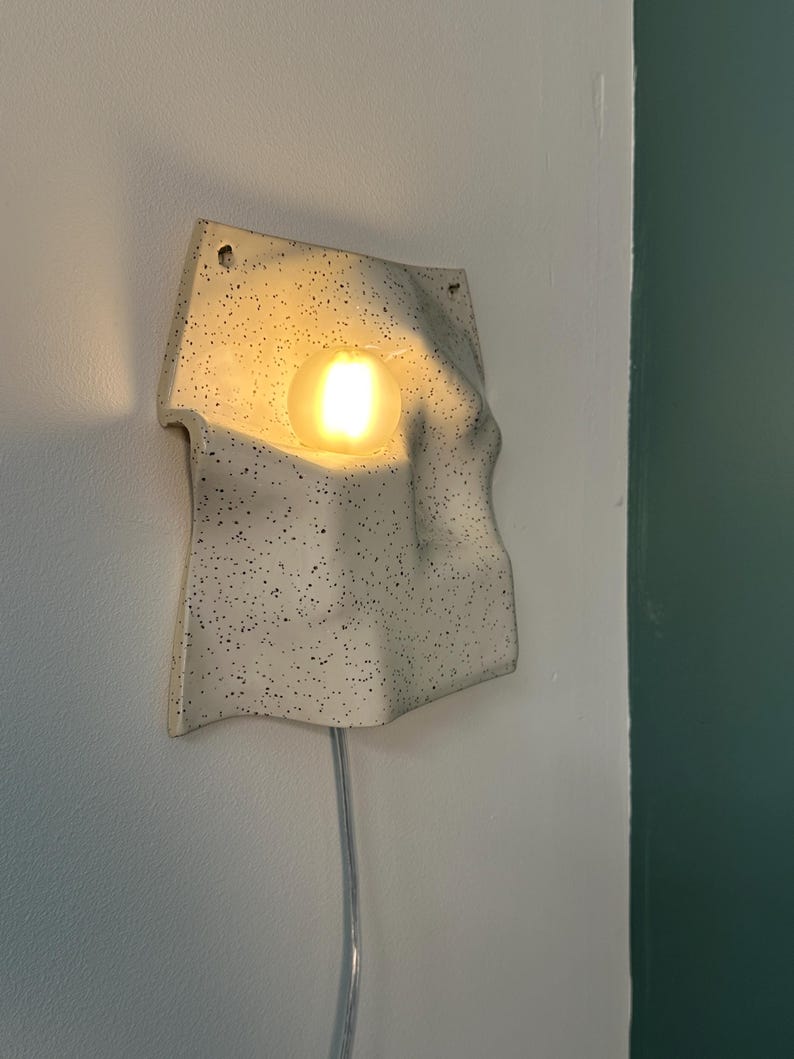 May include: A wall-mounted ceramic lamp with an off-white speckled finish and an irregular, wavy surface. The fixture features a central circular opening containing a glowing, warm-toned light bulb and includes a clear power cord extending downward.