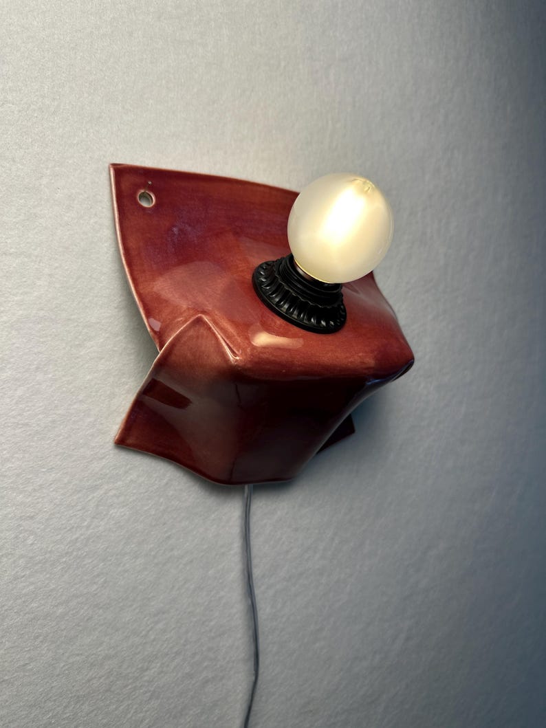 Puede incluir: Un aplique de pared con una base de cer&aacute;mica de color rojo oscuro brillante y forma org&aacute;nica plegada. Un casquillo negro sostiene una bombilla redonda de color blanco esmerilado. Un cable el&eacute;ctrico fino y claro cuelga de la l&aacute;mpara.