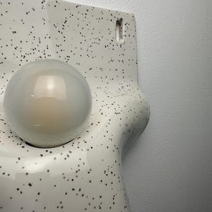Może przedstawiać: Kinkiet ścienny z błyszczącą białą ceramiczną podstawą w czarne kropki. Lampa ma zakrzywiony, organiczny kształt i zawiera pojedynczą, kulistą, matową żar&oacute;wkę umieszczoną w okrągłym otworze.