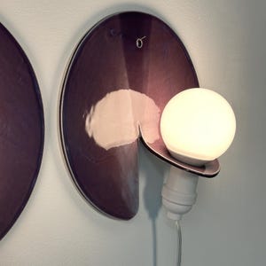 K&ouml;nnte beinhalten: Eine Wandleuchte mit einer kreisf&ouml;rmigen, dunkelvioletten Scheibe in Metallic-Optik und einem Ausschnitt. Eine wei&szlig;e Gl&uuml;hbirne sitzt in einer wei&szlig;en Fassung am unteren Rand der Scheibe, mit einem transparenten Stromkabel nach unten.