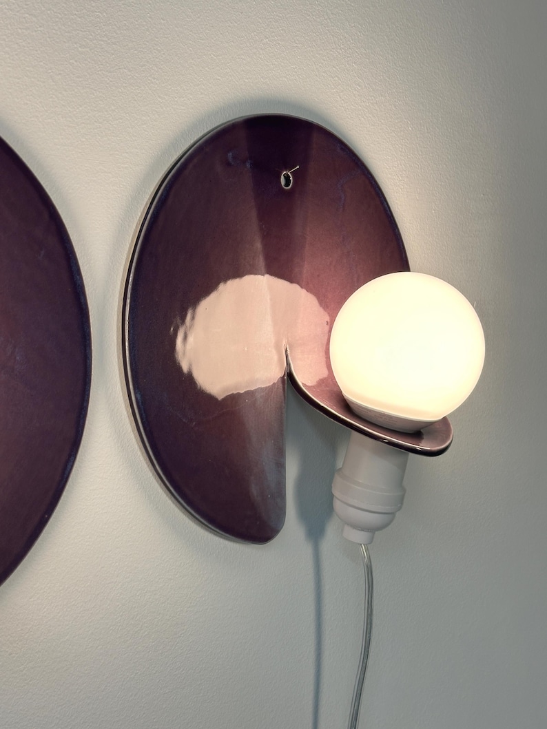 K&ouml;nnte beinhalten: Eine Wandleuchte mit einer kreisf&ouml;rmigen, dunkelvioletten Scheibe in Metallic-Optik und einem Ausschnitt. Eine wei&szlig;e Gl&uuml;hbirne sitzt in einer wei&szlig;en Fassung am unteren Rand der Scheibe, mit einem transparenten Stromkabel nach unten.