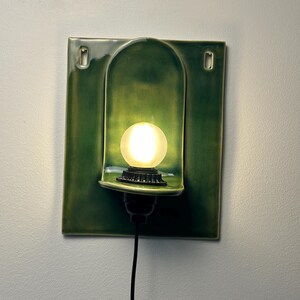 May include: A rectangular green ceramic wall sconce featuring a central arched niche that houses a glowing, frosted spherical light bulb. The fixture includes two mounting holes at the top corners and a black power cord extending from the base.
