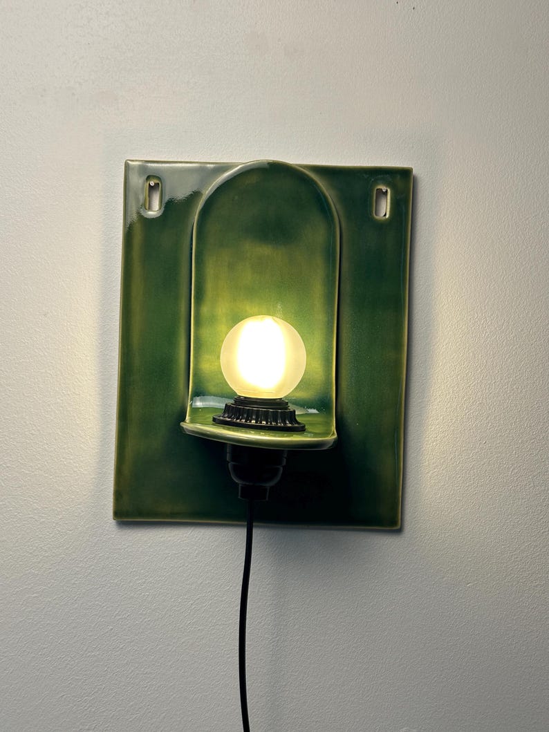 May include: A rectangular green ceramic wall sconce featuring a central arched niche that houses a glowing, frosted spherical light bulb. The fixture includes two mounting holes at the top corners and a black power cord extending from the base.