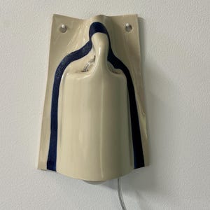 May include: A wall-mounted ceramic light fixture with a glossy cream-colored finish and a dark blue painted stripe accent. The structure features a curved, molded shape with two circular holes at the top for mounting and a clear electrical cord.