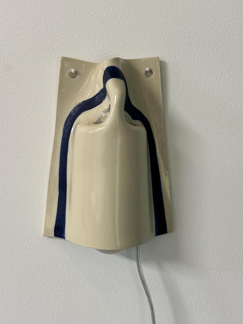 May include: A wall-mounted ceramic light fixture with a glossy cream-colored finish and a dark blue painted stripe accent. The structure features a curved, molded shape with two circular holes at the top for mounting and a clear electrical cord.