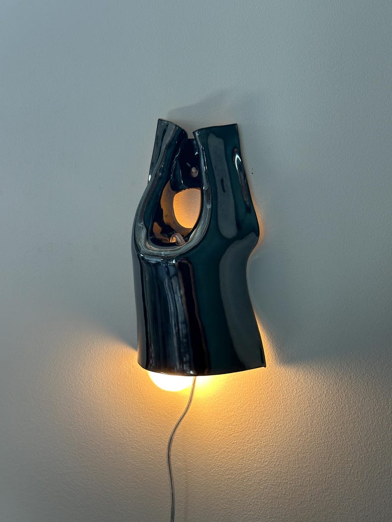 May include: A dark, glossy ceramic wall sconce with a smooth, curved, organic silhouette. The fixture features a central opening and emits a warm, soft glow from a light bulb positioned at the bottom, with a thin power cord extending downwards.