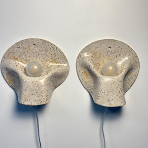 May include: A pair of wall sconces featuring a speckled off-white ceramic base with a wavy, organic shape. Each sconce has a central circular opening holding a frosted glass light bulb and a small hole at the top for mounting, with a power cord.