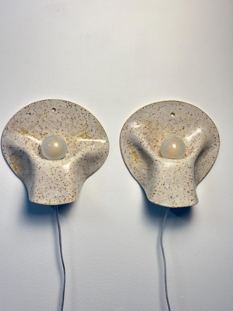 May include: A pair of wall sconces featuring a speckled off-white ceramic base with a wavy, organic shape. Each sconce has a central circular opening holding a frosted glass light bulb and a small hole at the top for mounting, with a power cord.