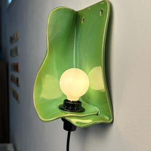 Pu&ograve; includere: Una lampada da parete con un paralume in ceramica verde brillante dalla forma organica e ondulata. L'apparecchio include una lampadina bianca rotonda e luminosa inserita in un portalampada nero, con un cavo di alimentazione nero.