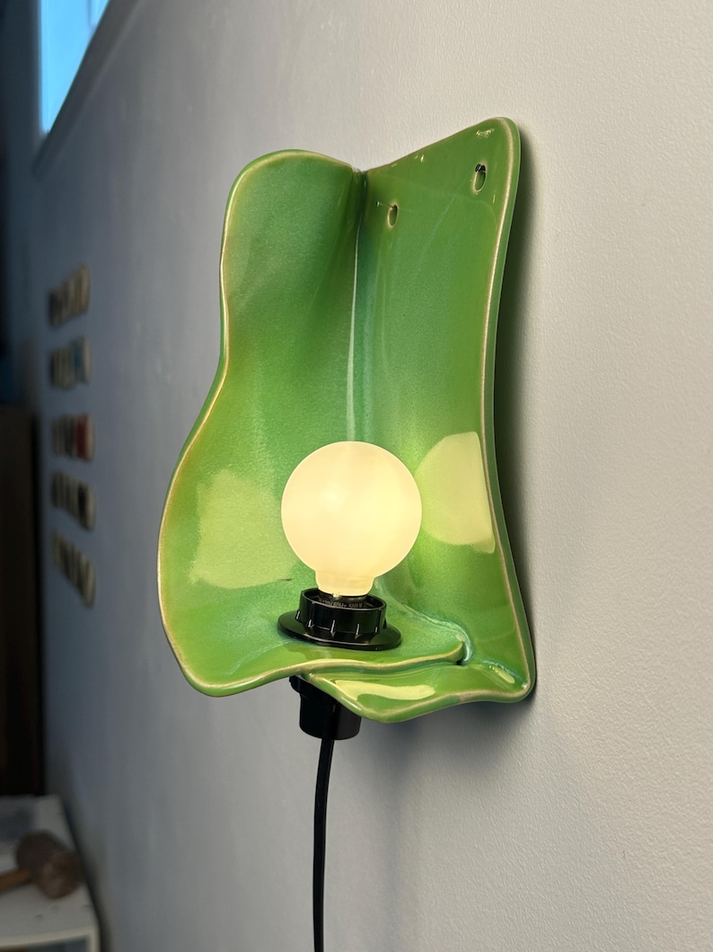 Pu&ograve; includere: Una lampada da parete con un paralume in ceramica verde brillante dalla forma organica e ondulata. L'apparecchio include una lampadina bianca rotonda e luminosa inserita in un portalampada nero, con un cavo di alimentazione nero.