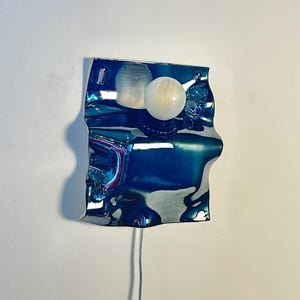 May include: A wall-mounted lamp featuring a reflective, wavy, deep blue metallic plate with a smooth, glossy finish. A single, spherical white glass light bulb is attached to the center of the plate, extending outward from the surface.