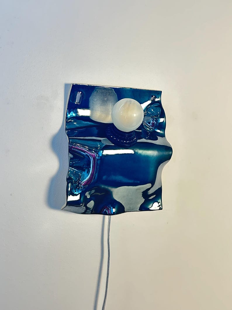 May include: A wall-mounted lamp featuring a reflective, wavy, deep blue metallic plate with a smooth, glossy finish. A single, spherical white glass light bulb is attached to the center of the plate, extending outward from the surface.