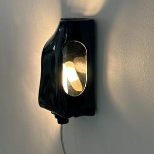 May include: A glossy black wall-mounted sconce with an organic, asymmetrical shape. The fixture features a central vertical oval cutout that reveals a warm, glowing light source, casting a soft illumination onto the surrounding wall surface.