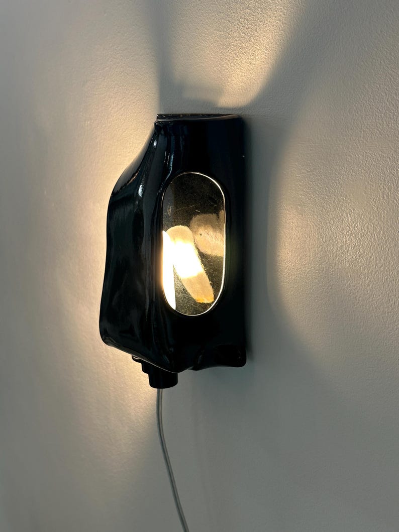 May include: A glossy black wall-mounted sconce with an organic, asymmetrical shape. The fixture features a central vertical oval cutout that reveals a warm, glowing light source, casting a soft illumination onto the surrounding wall surface.