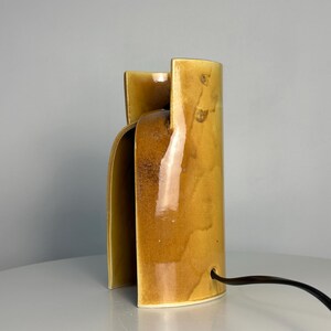Puede incluir: Una l&aacute;mpara de mesa cil&iacute;ndrica de cer&aacute;mica con un esmalte amarillo mostaza brillante y un dise&ntilde;o de corte vertical. La l&aacute;mpara presenta una superficie lisa y curva con sutiles variaciones tonales en el acabado y un cable el&eacute;ctrico negro.