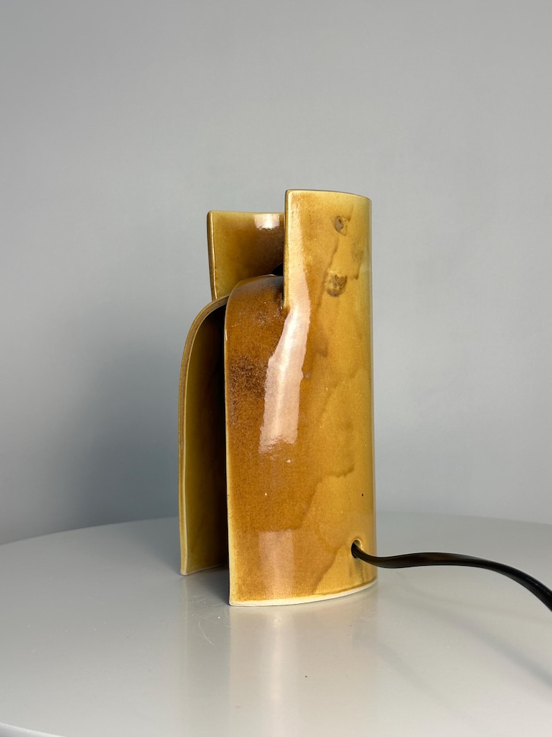 Puede incluir: Una l&aacute;mpara de mesa cil&iacute;ndrica de cer&aacute;mica con un esmalte amarillo mostaza brillante y un dise&ntilde;o de corte vertical. La l&aacute;mpara presenta una superficie lisa y curva con sutiles variaciones tonales en el acabado y un cable el&eacute;ctrico negro.