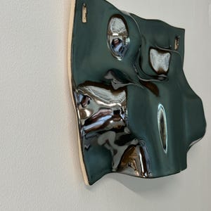 Pu&ograve; includere: Una scultura da parete in ceramica con una finitura lucida color verde ottanio scuro e una superficie ondulata altamente riflettente. L'oggetto presenta curve e rientranze organiche irregolari, con due piccole aperture rettangolari in alto.