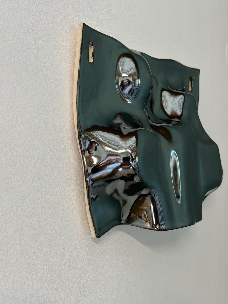 Pu&ograve; includere: Una scultura da parete in ceramica con una finitura lucida color verde ottanio scuro e una superficie ondulata altamente riflettente. L'oggetto presenta curve e rientranze organiche irregolari, con due piccole aperture rettangolari in alto.