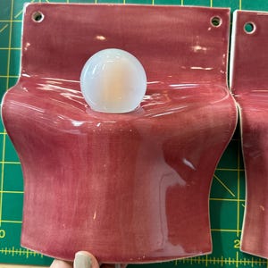 May include: A wall-mounted ceramic light fixture with a glossy, deep rose-colored glaze. The piece features a curved, wave-like form with two circular holes at the top for mounting and a central opening holding a small, round, frosted white light bulb.