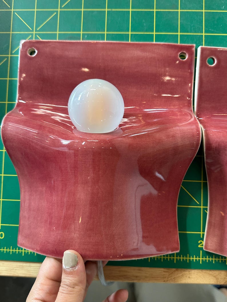 May include: A wall-mounted ceramic light fixture with a glossy, deep rose-colored glaze. The piece features a curved, wave-like form with two circular holes at the top for mounting and a central opening holding a small, round, frosted white light bulb.