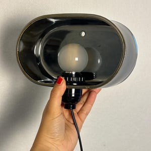 K&ouml;nnte beinhalten: Eine schwarze und bronzefarbene Wandleuchte mit einem ovalen Schirm. Die Leuchte hat eine Gl&uuml;hbirne und ein schwarzes Kabel. Der Schirm hat eine gl&auml;nzende Oberfl&auml;che und eine bronzefarbene Einfassung. Die Leuchte wird von einer Person gehalten.