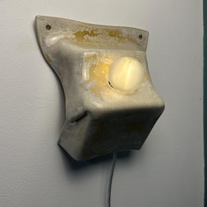May include: A wall-mounted light fixture with a textured, off-white, and translucent resin-like body. The structure features a flared top with two mounting holes and a central circular opening containing a glowing, spherical light bulb.