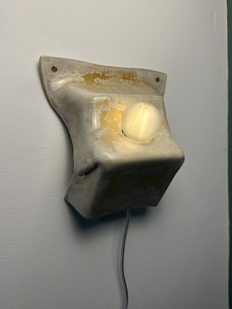 May include: A wall-mounted light fixture with a textured, off-white, and translucent resin-like body. The structure features a flared top with two mounting holes and a central circular opening containing a glowing, spherical light bulb.